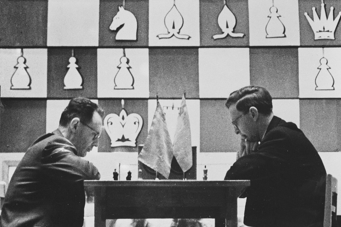 Black-and-white photo: two men in glasses bent over a chessboard on a stage, a giant demonstration board with pieces behind them; Botvinnik on the left, Smyslov on the right