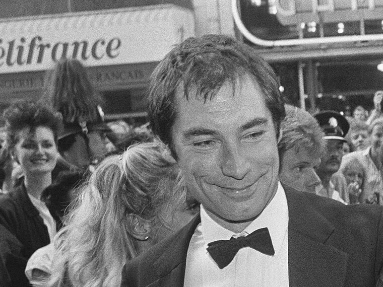 Timothy Dalton in a tuxedo and bow tie, smiling outside a cinema at a premiere, with a crowd behind him; black-and-white photo
