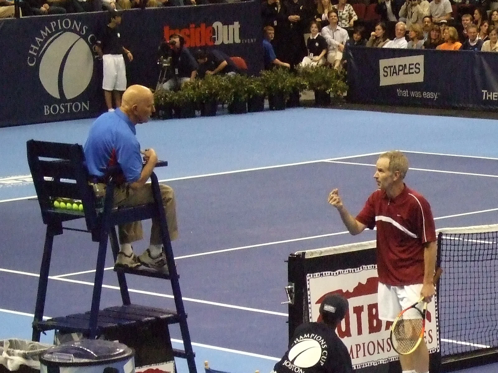 John McEnroe in a red shirt, racket in one hand, gestures at an umpire sitting in a high chair beside the court
