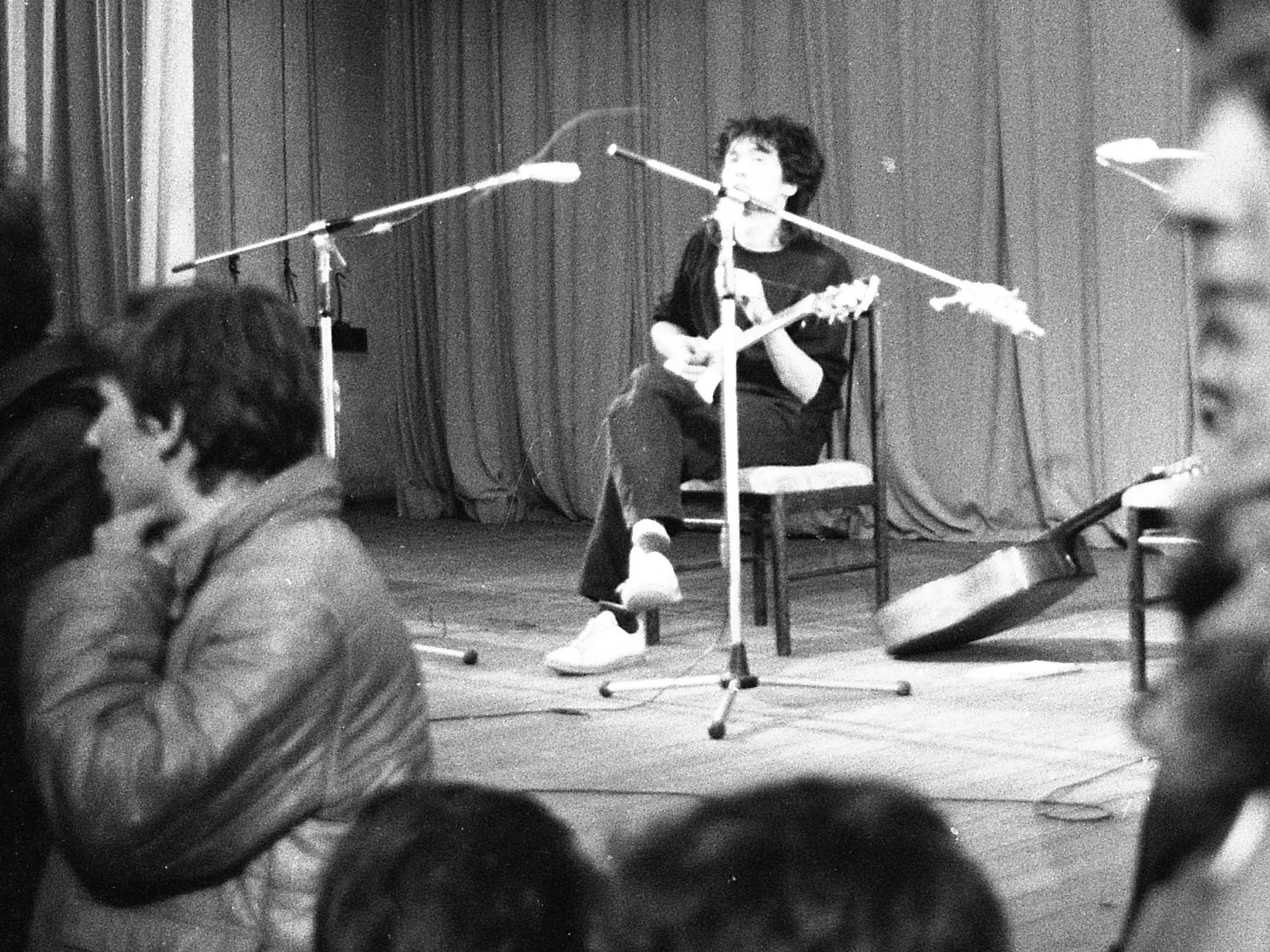 Viktor Tsoi sits on a chair on a small stage with an acoustic guitar between two microphone stands, with the audience in the foreground