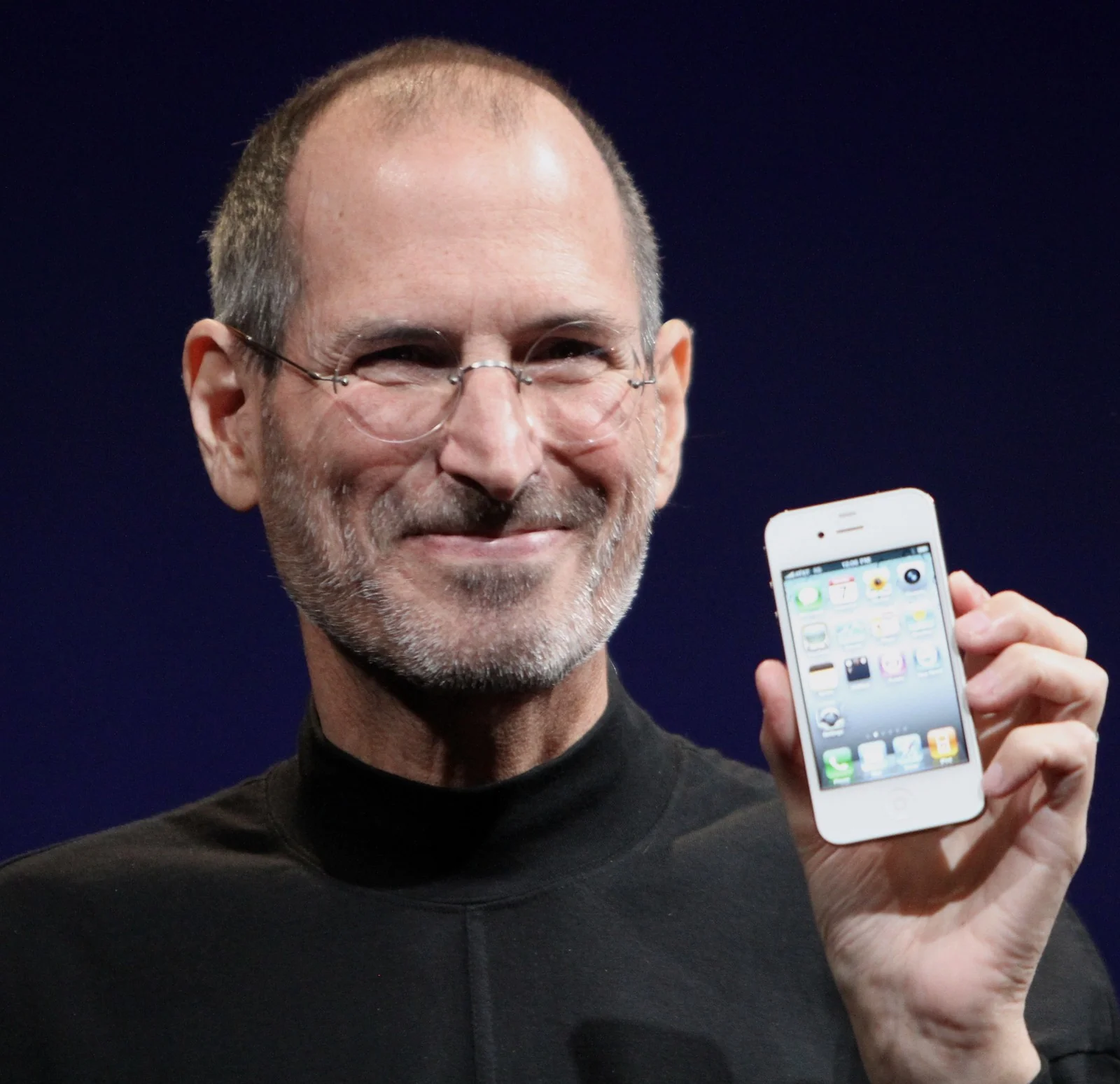 Steve Jobs in a black turtleneck and glasses smiles and holds up a white smartphone