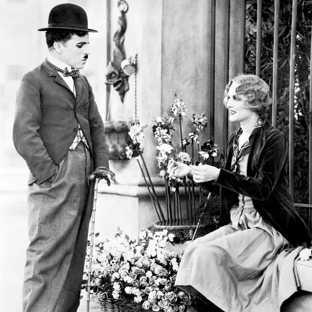 Charlie Chaplin in a bowler hat, holding a cane, stands before a young woman sitting by a railing next to a basket of flowers