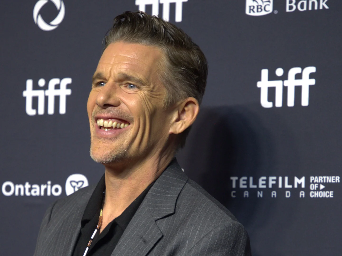 Ethan Hawke, smiling, in a gray jacket in front of a wall of festival logos