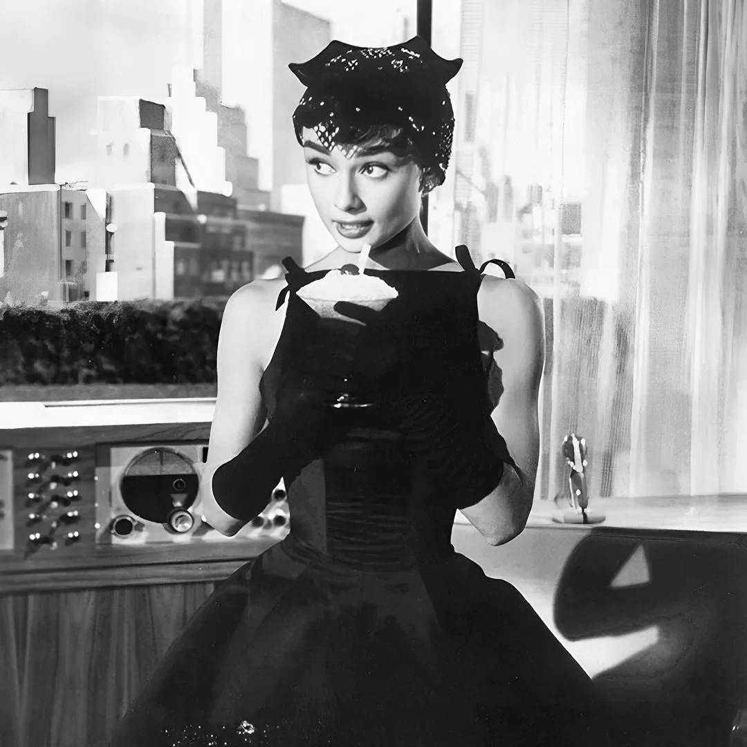 Audrey Hepburn in a black dress with bows on the shoulders, long gloves and a small hat, holding a glass in front of a window with a city view